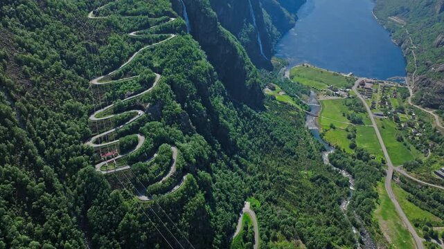 Close aerial view of Trollstigen hairpin bends surrounded by dense green forest. Iconic Norwegian mountain road appears dramatic and isolated on a sunny day.