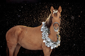 A brown horse stands gracefully against a dark background with falling snowflakes. The horse wears a festive Christmas wreath made of frosted branches and silver and gold ornaments around its neck