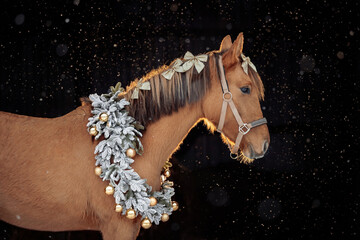 A brown horse stands gracefully against a dark background with falling snowflakes. The horse wears a festive Christmas wreath made of frosted branches and silver and gold ornaments around its neck