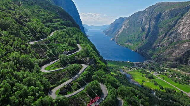 Aerial view of Trollstigen road above Geirangerfjord in Norway. Drone footage shows dramatic hairpin turns winding through green mountains toward the deep blue fjord on a clear summer day..
