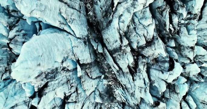 Top-down drone view of fractured blue glacier with dark volcanic ash bands and narrow crevasses, creating an abstract natural background.