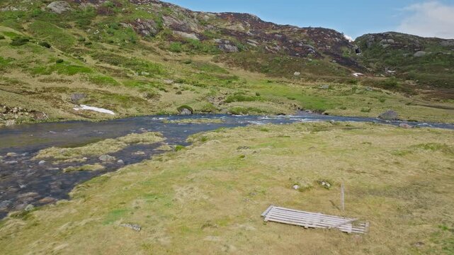 A calm stretch of mountain river curves through open grassland in Norway. Red wooden Norwegian house surrounded by rolling green hills, scattered rocks, and patches of snow beneath a bright, clear sky