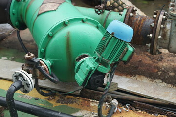 Close-up view of a robust industrial pump system with a green metallic body and an electric motor, connected by various pipes and valves, indicating essential process infrastructure