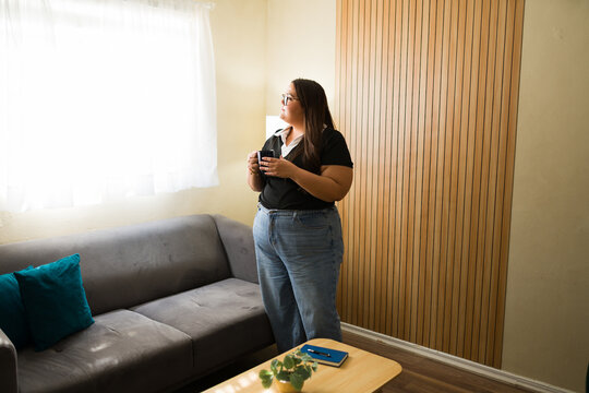 Pensive woman standing in living room enjoying coffee and contemplating while looking out window
