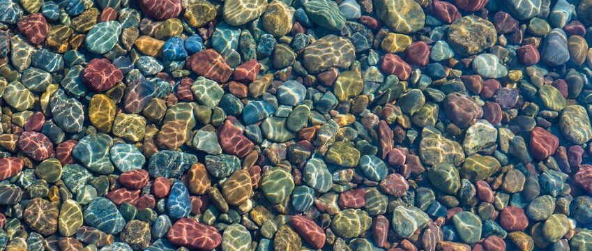An underwater scene showcases a mesmerizing collection of smooth, colorful river rocks, each glistening under the transparent water, creating a tranquil display of natural beauty.