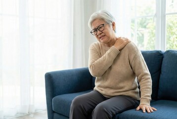 An elderly woman experiences shoulder pain while sitting on a couch in a bright living room with natural light.