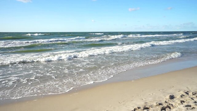 A windy seaside scene with waves breaking along a sandy beach beneath a bright blue sky with drifting clouds.