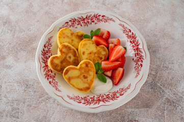 Heart-shaped cottage cheese pancakes or syrnyky with sour cream and fresh strawberries on a plate on a concrete background. A breakfast for Valentine's Day or Mother's Day. Top view