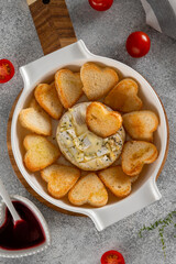 Baked Camembert cheese with garlic and thyme in a white ceramic baking dish, served with heart-shaped toasted bread. Romantic appetizer or snack for Valentine's Day or date night. Top view.