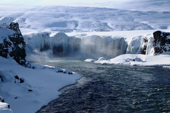 Partially frozen waterfall cascading through snowy terrain with mist and icy cliffs in winter landscape