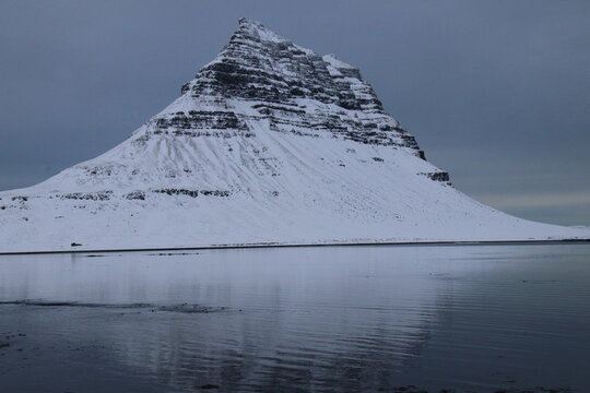 Snow-covered conical mountain reflected in calm water under overcast sky in remote landscape
