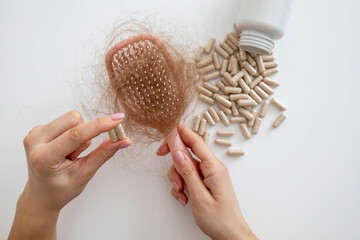 Woman holding hair loss vitamins, hairbrush with fallen hair and supplement capsules on white background