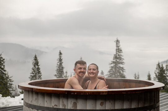 A couple in love enjoying hot tub outdoors. Winter holidays in snowy mountains. Family relax in thermal spa open air. Man and woman swims in hot bath while resting. Joyful young man hugging a woman.