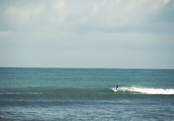 A surfer on his board riding a long, blue, crystal-clear wave; with the sea and sky in the background.