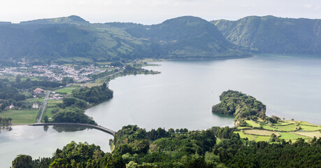 Volcanic caldera lake in Sete Cidades, Sao Miguel, Azores. Lush green hills and mountains surrounding the tranquil blue water. Scenic landscape of Portugal with small islands in the lagoon.