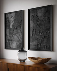 Two textured black framed artworks hang above a wooden cabinet with a glass vase and bowl