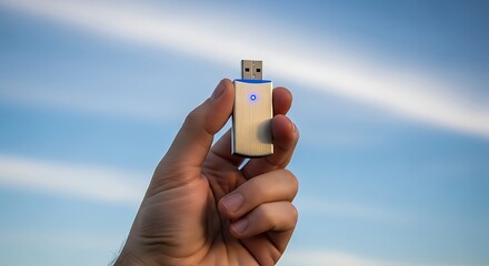 A hand holding a silver and blue usb flash drive against a sky