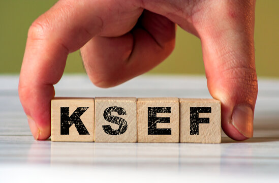 A hand holds wooden blocks with the letters KSEF prominently displayed on a light wooden surface. The scene suggests themes of business, regulation, and finance.