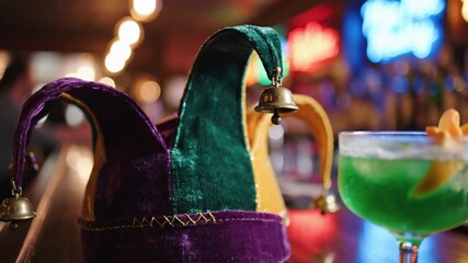 A colorful velvet jesters hat with small bells rests on a wooden bar next to a vibrant green cocktail garnished with citrus peel against a blurred background of warm bar lights