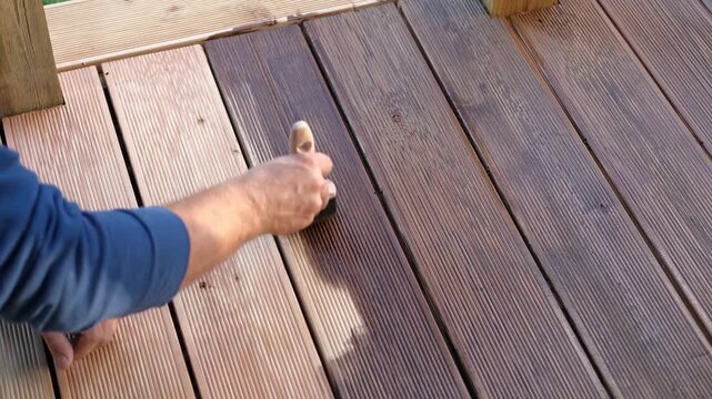 Brown oil for protection of wood by staining. Painting a larch for protecting and pests. Woodworker applying finish to terrace deck board. Repairman varnishing with brush wooden plank floor surface.