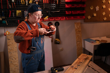 professional female carpenter works with wood using carpentry tools in her garage. She stands holding a phone and takes photos of the workpieces. Conceptualization of profession, art, and hobby.