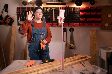 A professional female carpenter works with wood using carpentry tools in her garage. She makes a video call using a smartphone. Concept of profession, art, and hobby