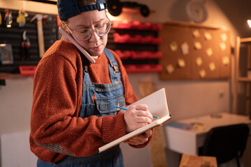 A professional carpenter works with wood using carpentry tools in her garage, taking orders over the phone and writing them down in a notebook. Craft, creative small business