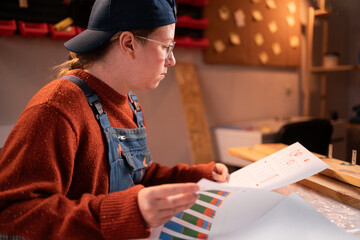 A professional carpenter works with wood using carpentry tools in her garage. She holds sheets of paper with diagrams and studies them. Craft, creative small business.