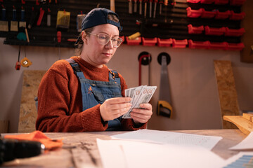 A professional carpenter works with wood using carpentry tools in her garage. She sits at a table, holding dollars in her hands, and counts money. Craft, creative small business.