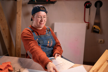 professional female carpenter works with wood using carpentry tools in her garage. She sits, holds sheets of paper with graphs, and studies the material. Concept of profession, art, and hobby.