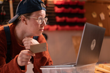 professional female carpenter works with wood using carpentry tools in her garage. She holds a notebook and shops online using a laptop. Concept of profession, art, and hobby.