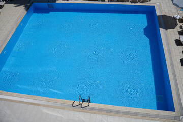 Clear blue water in square pool, sparkling in midday sun, with tiled liner and stainless steel staircase. Relaxation concept, summer fun
