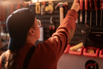 female carpenter works in her garage, using professional woodworking tools. Handicrafts, creativity, and carpentry. She stands near a tool stand and picks up something.