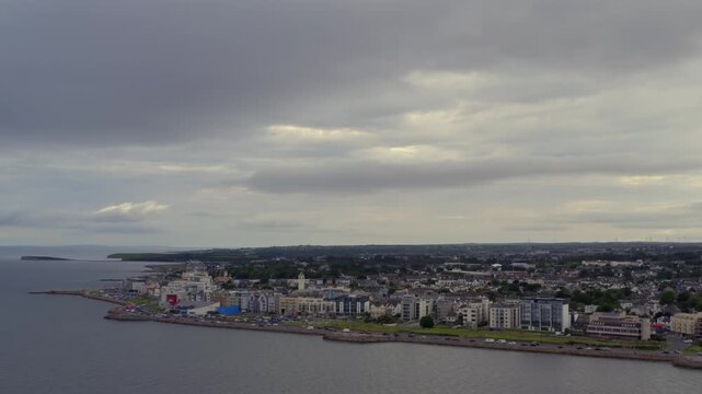 Aerial dolly shot approaching Salthill village from Galway Bay beneath a dramatic, overcast sky.