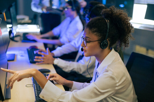 Professional young female operator wearing headset pointing at computer screen while working with diverse team in a high-tech call center or mission control room during night shift operations