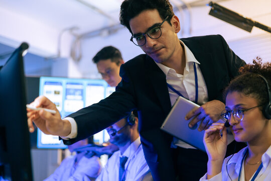 Dedicated professional team collaborates in modern tech office. Manager points at monitor screen while female colleague in headset provides customer support and digital data analysis service - Powered by Adobe