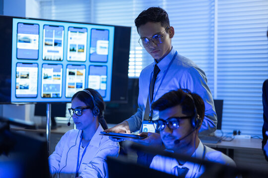 Professional IT support team working in a high-tech control room. Focused male supervisor holding a digital tablet while monitoring mobile app interface designs on a screen with technicians - Powered by Adobe