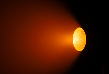 Close up of a studio spotlight emitting warm orange light with a textured reflector in a dark surrounding space. Lighting equipment backstage.