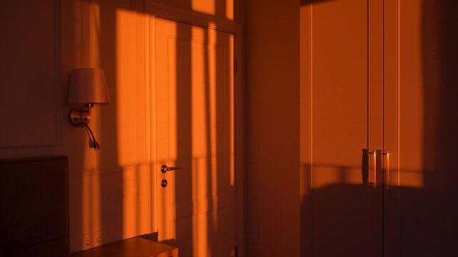 Beautiful golden hour sunlight casting long, moving shadows across a bedroom wall and furniture. Warm, orange light from the setting sun illuminates the tranquil and cozy room interior