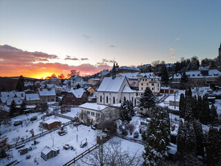 peaceful winter scene of a quaint village blanketed in snow, with a church at its center and a glowing sunset casting warm light over the quiet rooftops, Hartenstein - Germany
