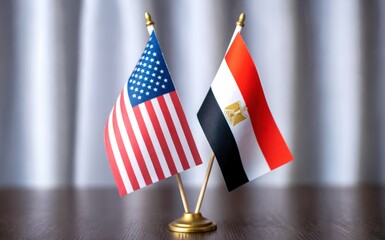 Two miniature national flags prominently displayed side by side on a polished dark brown wooden surface an American flag and an Egyptian flag standing tall with a subtly textured light gray background