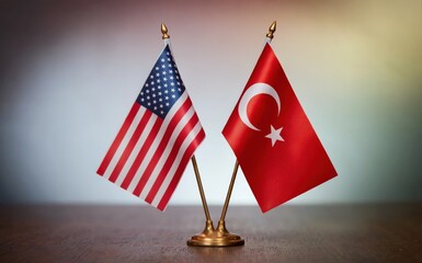 Two small national flags representing the United States of America and Turkey stand upright side by side on a polished wooden table against a softly blurred neutral gradient background illuminated by 