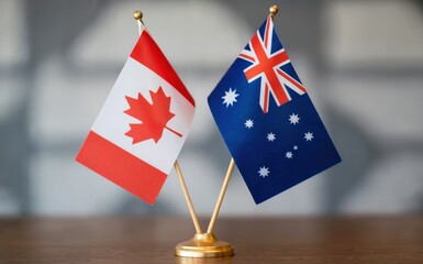 Two small miniature flags representing Canada and Australia stand upright side by side on a polished wooden desk The light grey backdrop provides a neutral setting Soft even lighting illuminates the s