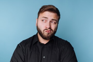 Obraz premium Man with a beard in a black shirt making a skeptical expression against a blue background.