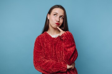 A young woman in a red sweater, looking thoughtful with a finger on her chin, against a light blue...