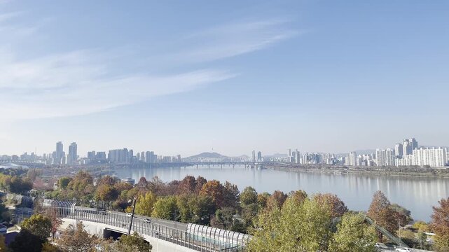 Wide panoramic view of Seoul city skyline along the Han River during autumn season. Calm river, colorful fall foliage, modern urban buildings and clear blue sky create a peaceful city landscape in Sou