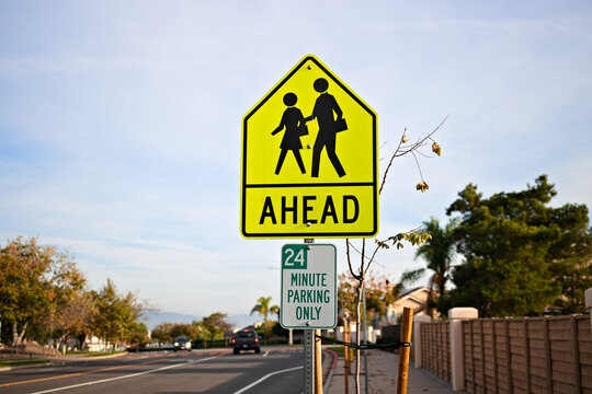 Yellow pedestrian crossing sign warns drivers of a school zone ahead along a quiet suburban street under clear daylight.