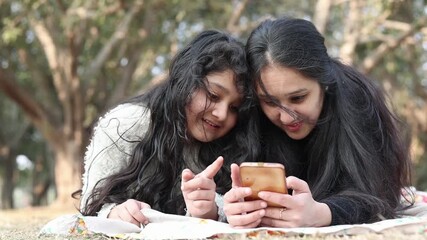 Closeup of Indian mother and daughter lying down in a park during a picnic while looking on camera through a smartphone. Happy family bonding moment with smiles and outdoor joy.