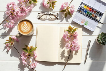 Flat lay of an artist's workspace with a blank open notebook, cherry blossoms, a cup of latte art, watercolor paints, and glasses on a white wooden table. Elegant spring mockup for design.