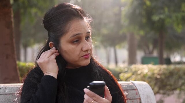 Closeup of an Indian woman wearing an earpod in her ear while sitting in a park and looking on camera. Calm outdoor lifestyle moment showing technology, focus, and modern daily life.